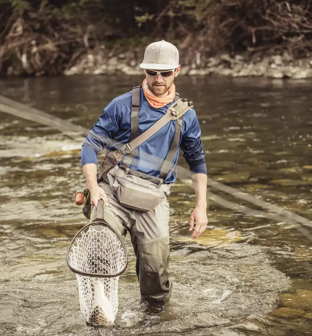 Man fishing in a river with gear, wearing sunglasses and a hat.