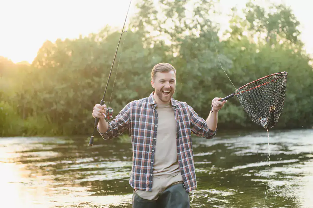 Fishing enthusiast with a fishing rod and net at the riverbank during sunset.